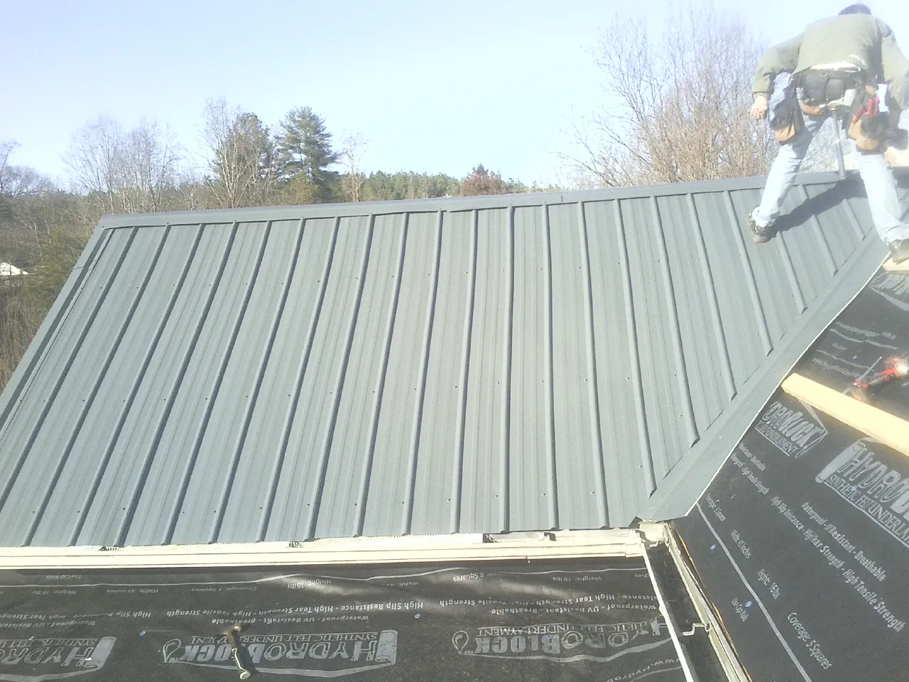 Crew installing dark green standing seam panels over synthetic underlayment in Tamassee