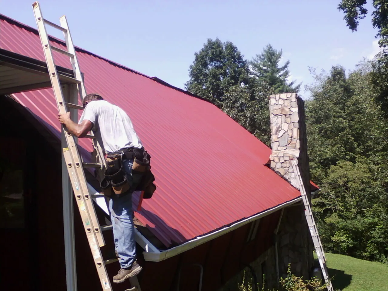 Roofer on a ladder beside a red metal roof and stone chimney — Soffit & Fascia Repair by Roofing Brothers in Tamassee